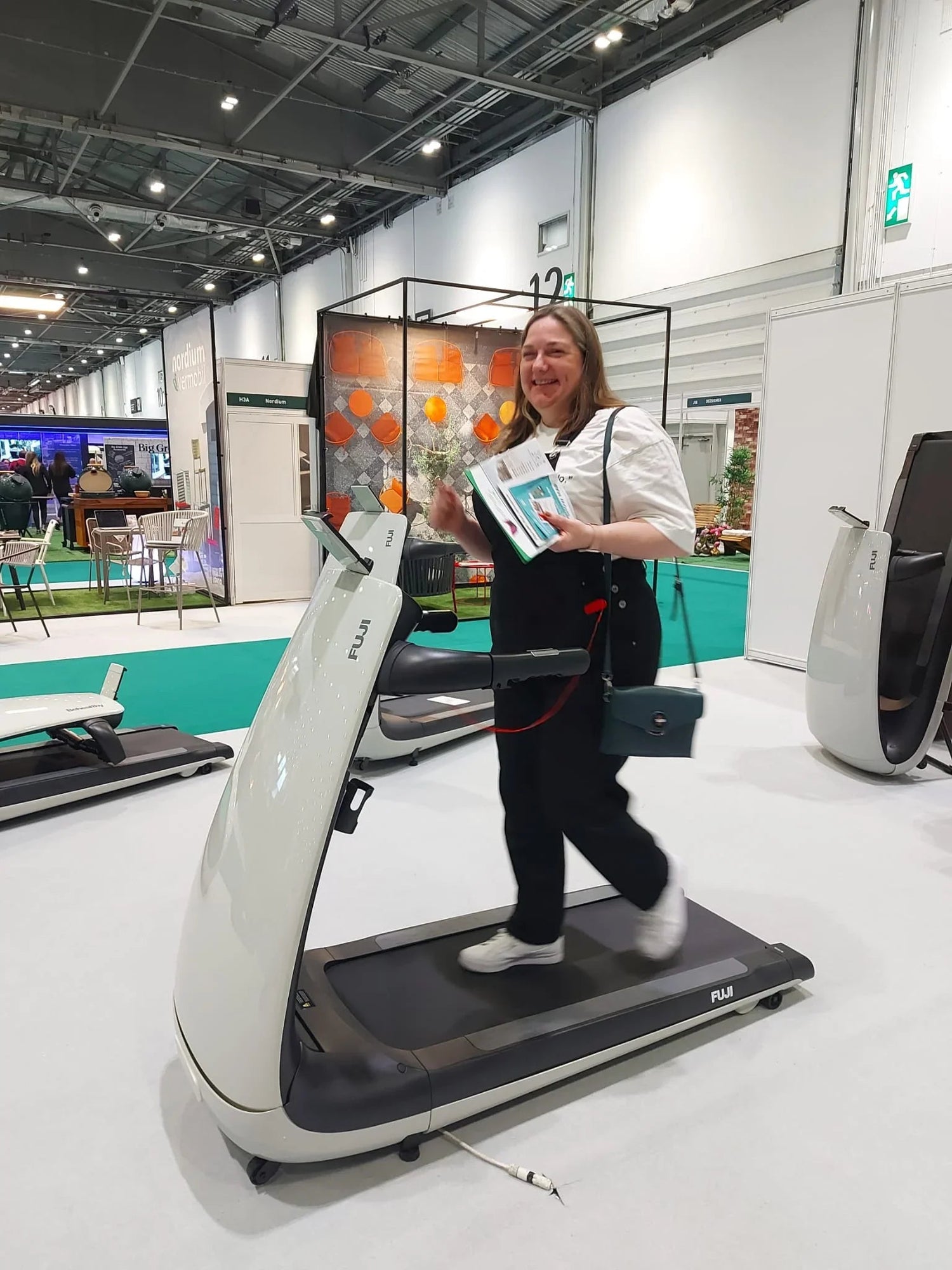 Woman walking on FUJI treadmill at indoor trade show, smiling and holding brochures